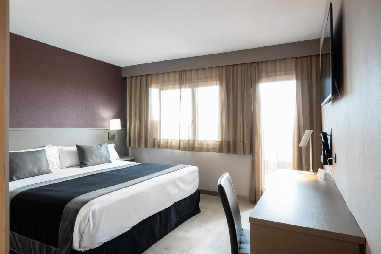 A hotel room with a bed, a desk, a chair, a TV, and a window with curtains.