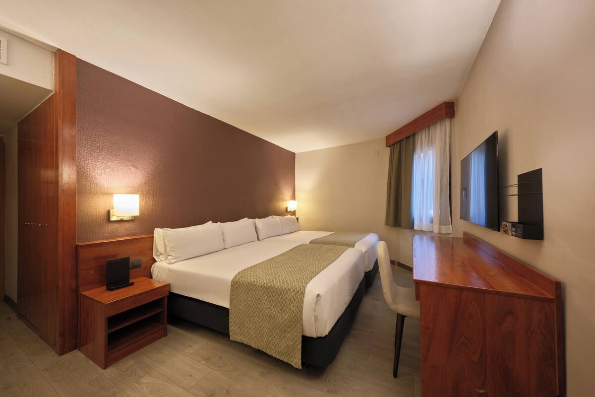 Habitación de hotel con una cama grande, mesitas de noche, un escritorio, una silla, un televisor y un ventanal con cortinas.