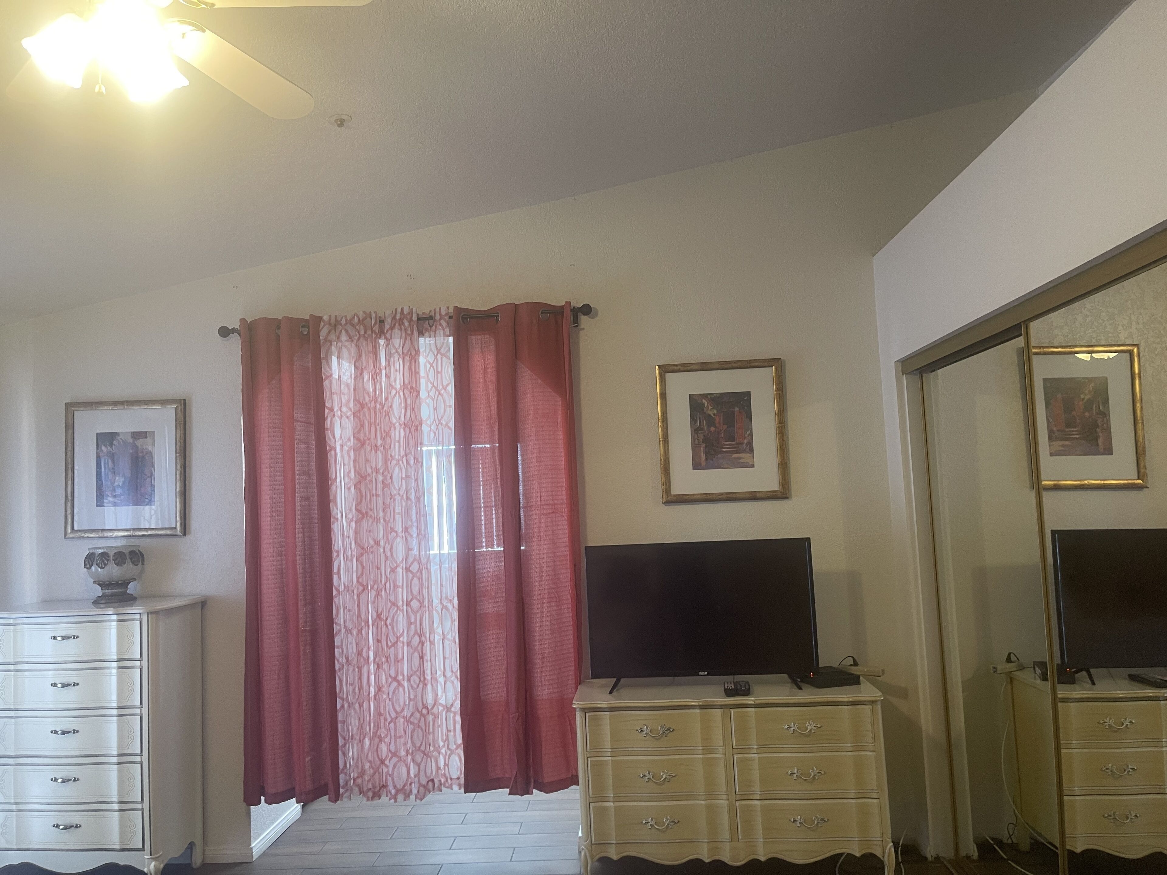 A room with a ceiling fan, a TV on a dresser, a large window with red curtains, and a mirror with a picture frame.
