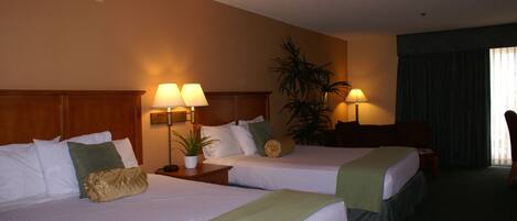 A hotel room with two beds, a wooden headboard, a nightstand with a lamp, a potted plant, and a large window with curtains.