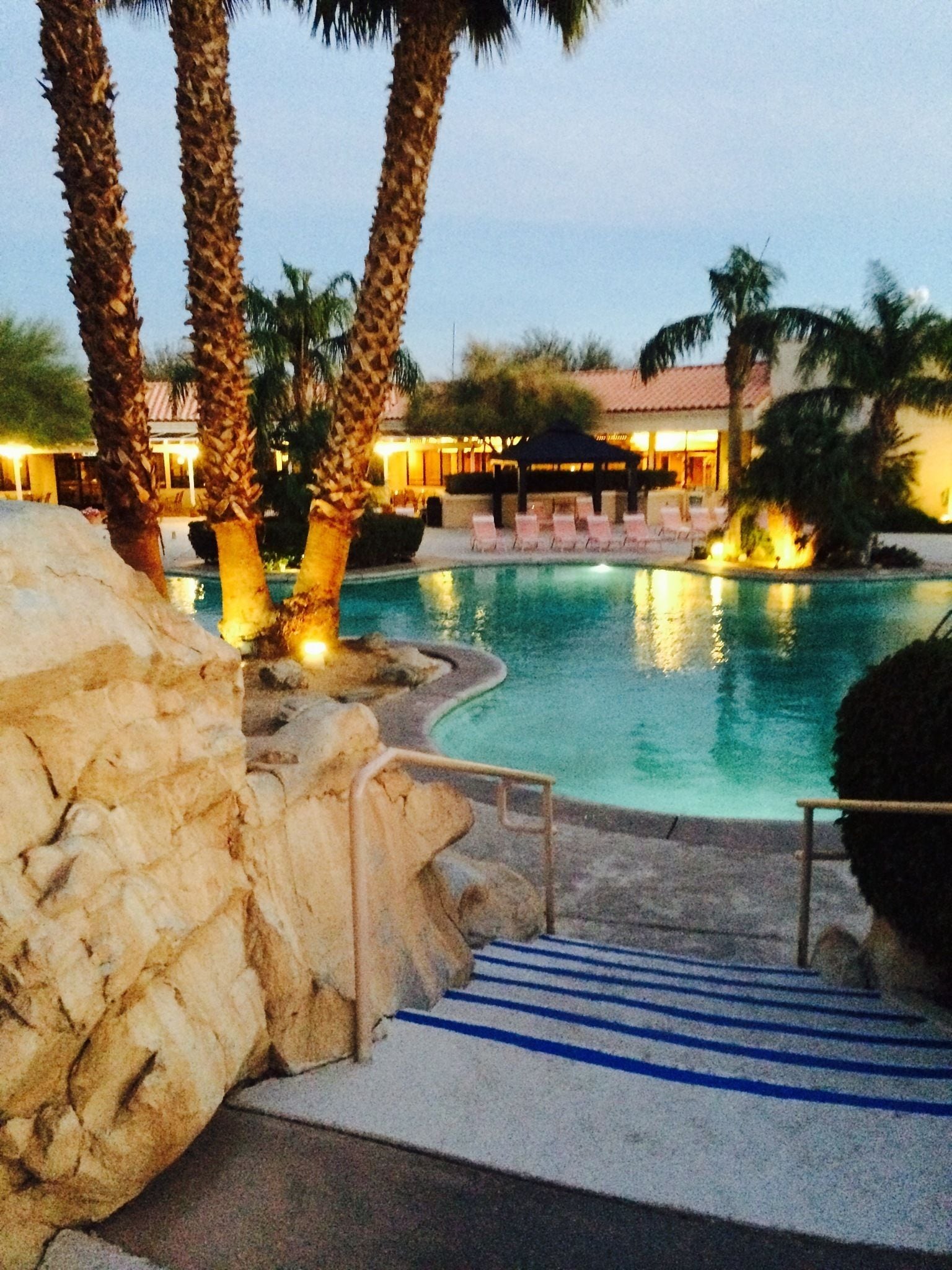 A swimming pool with palm trees, lounge chairs, and a building in the background.