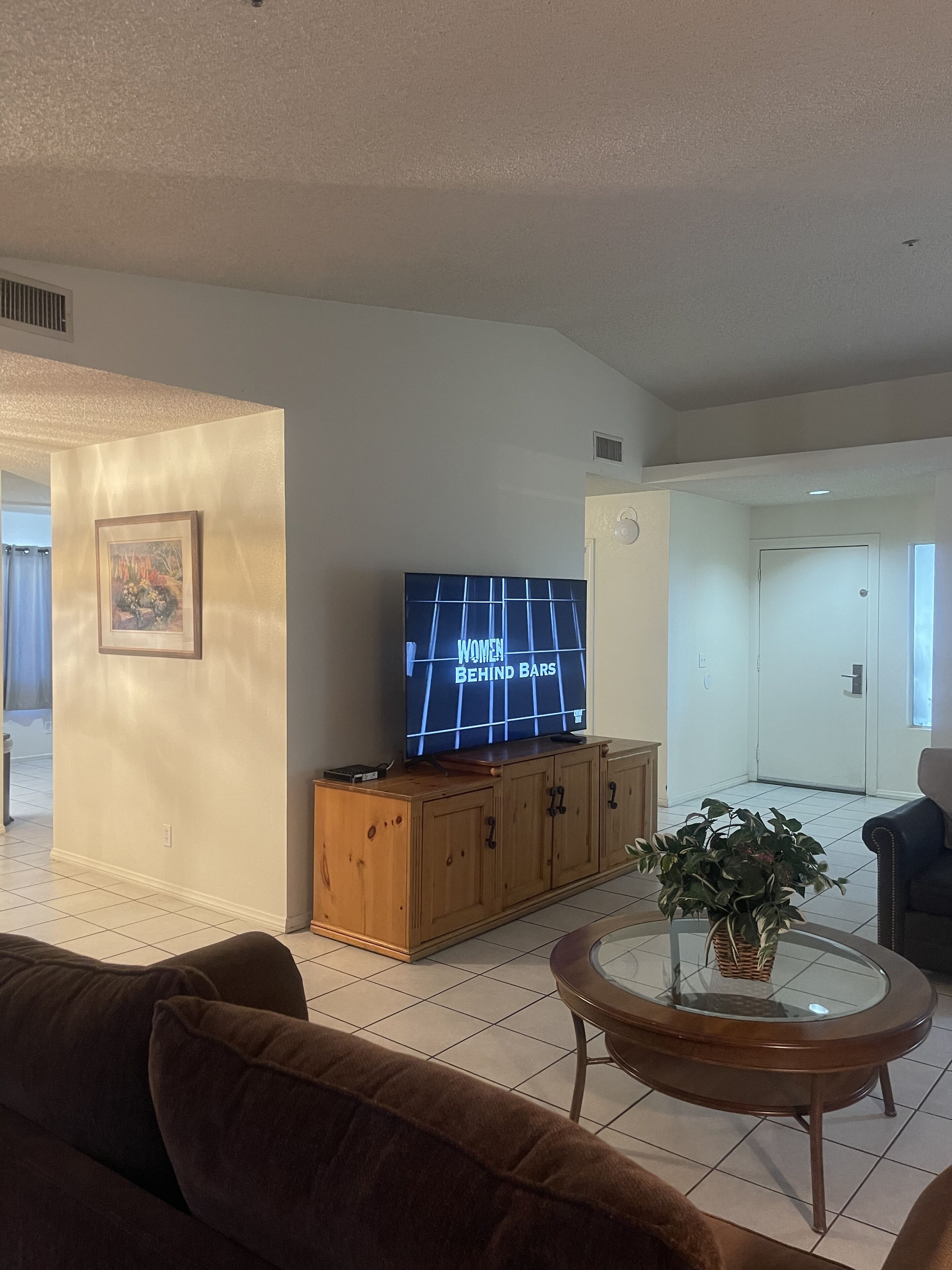 Una sala de estar con una televisión, un mueble de madera, una mesa de centro de vidrio y un sofá marrón.