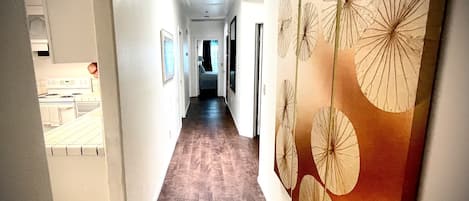 A hallway with a wooden floor, white walls, and a ceiling light. There are framed pictures on the walls and a door at the end of the hallway.