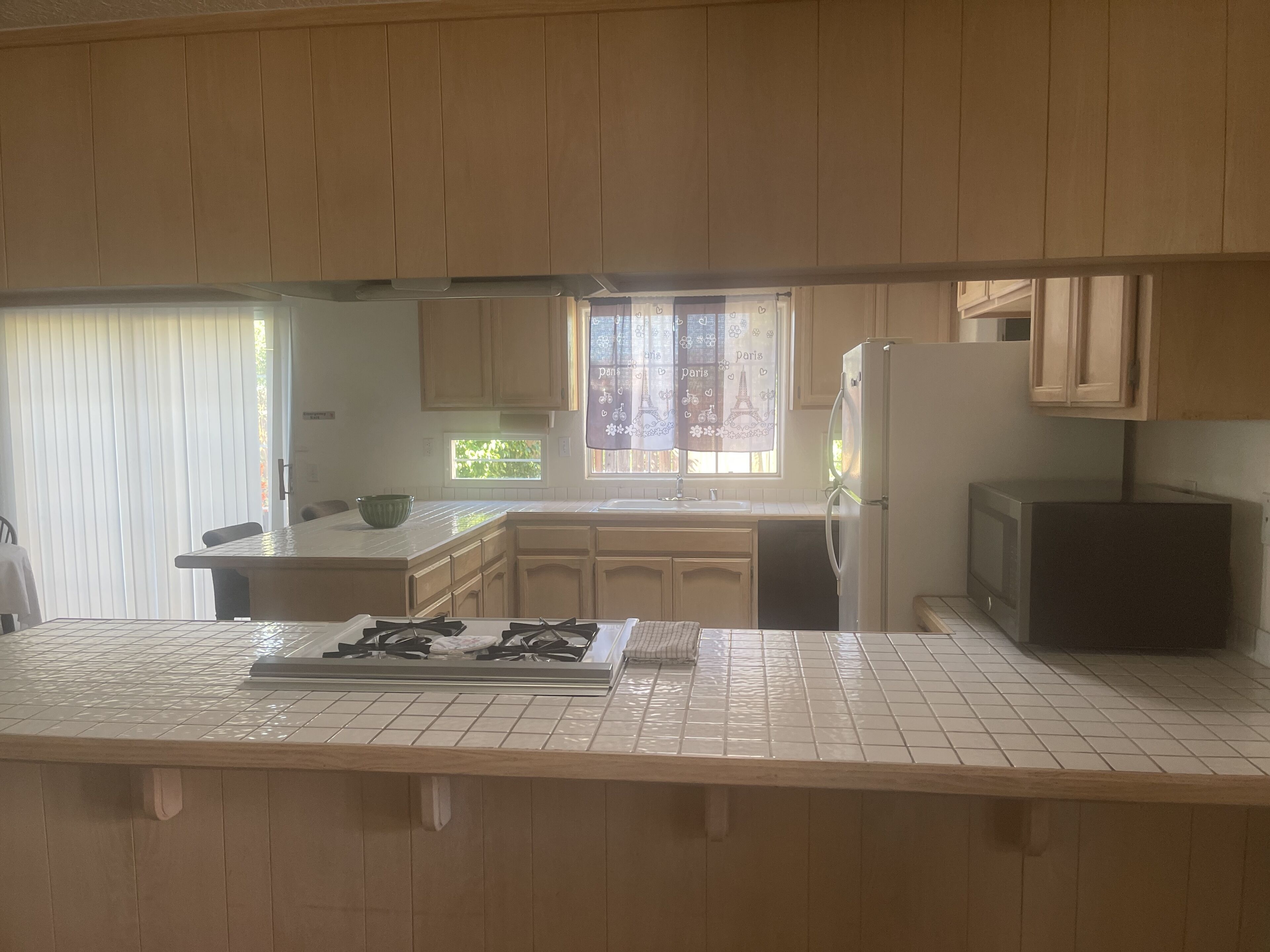 Een keuken met houten kasten, een gasfornuis, een koelkast en een raam met uitzicht op groen.