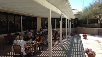 Eine Terrasse im Freien mit Tischen, Stühlen, großen Fenstern und einer Pergola.