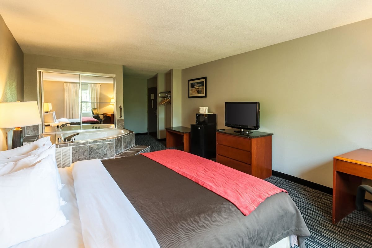 A hotel room with a bed, a desk, a television, a desk lamp, a coffee maker, and a bathroom with a bathtub and mirror.