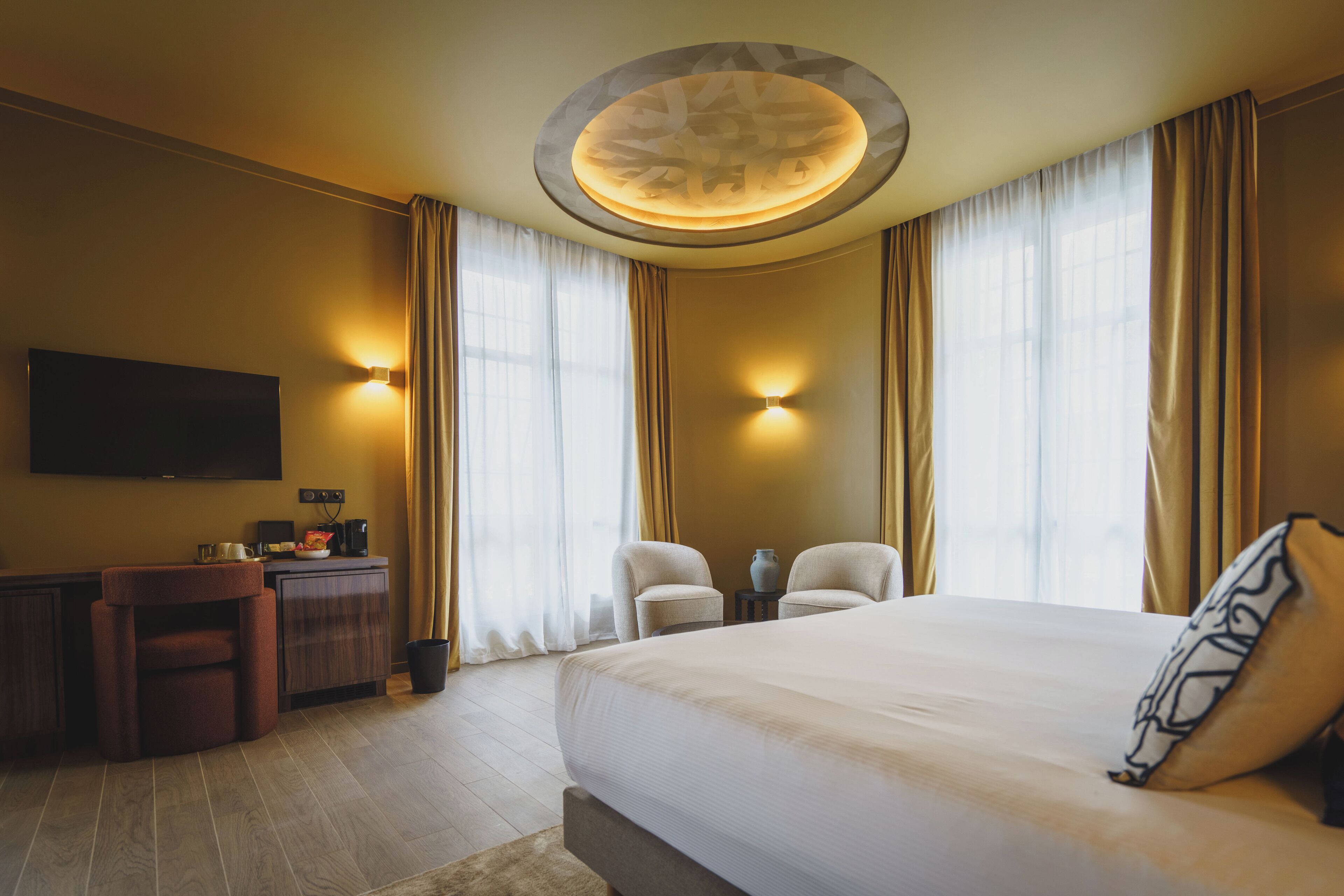 A hotel room with a large bed, a desk, two chairs, a flat-screen TV, and a ceiling light fixture.