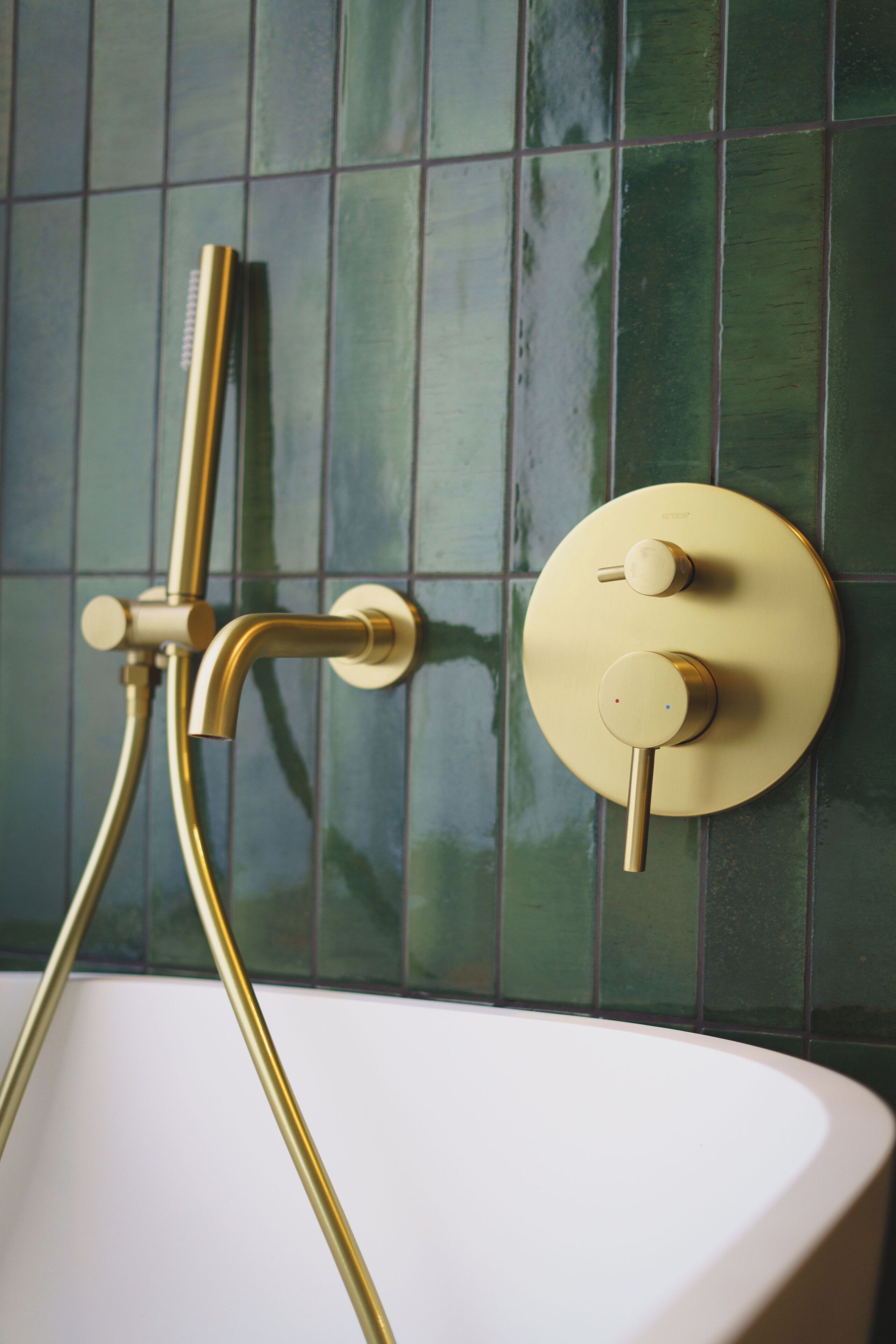 Une salle de bain avec une robinetterie de douche dorée et une baignoire blanche, le tout sur un mur recouvert de carreaux verts.