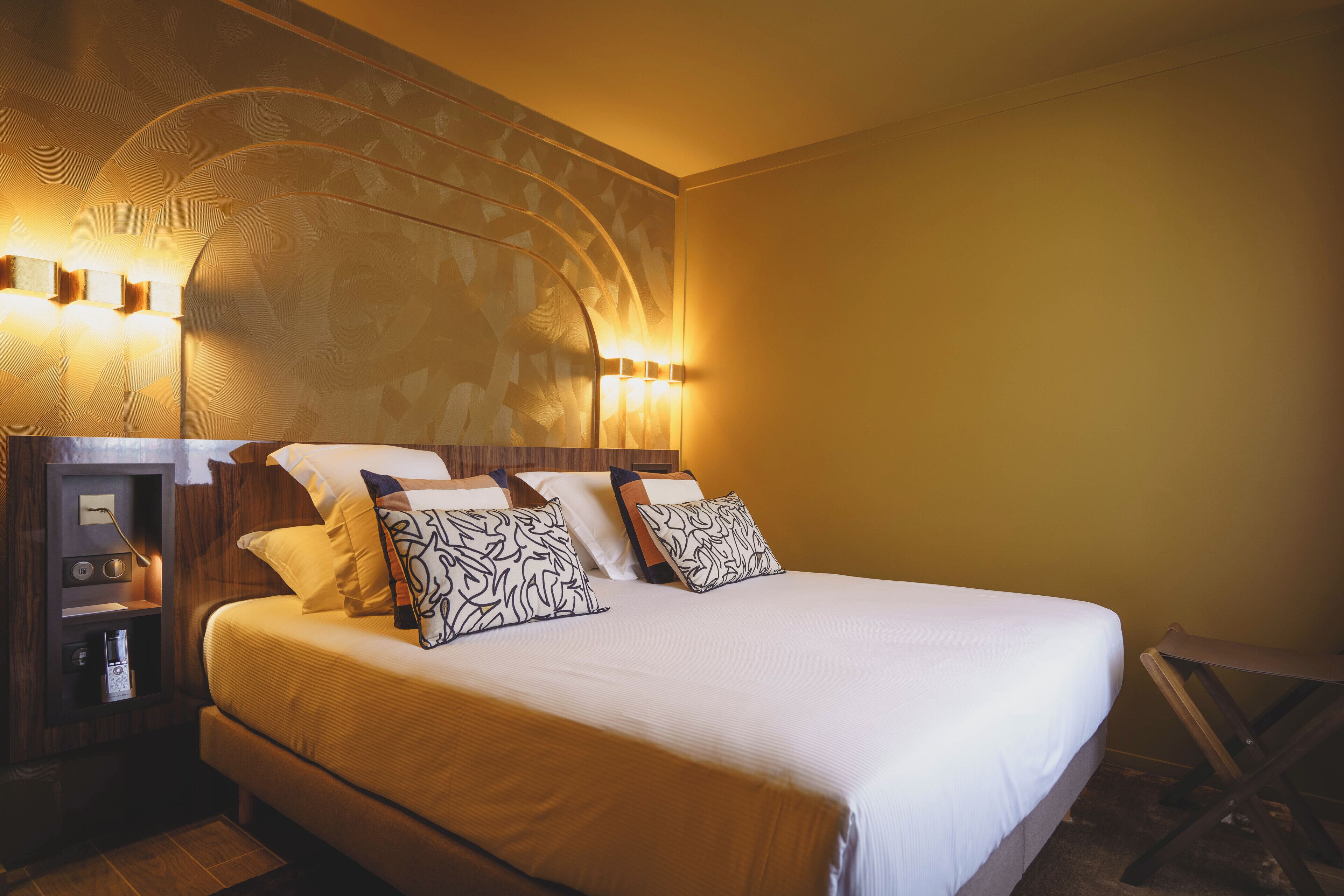 A neatly made bed with white bedding and patterned pillows, a wooden headboard with built-in lighting, and a small bedside table with a lamp.