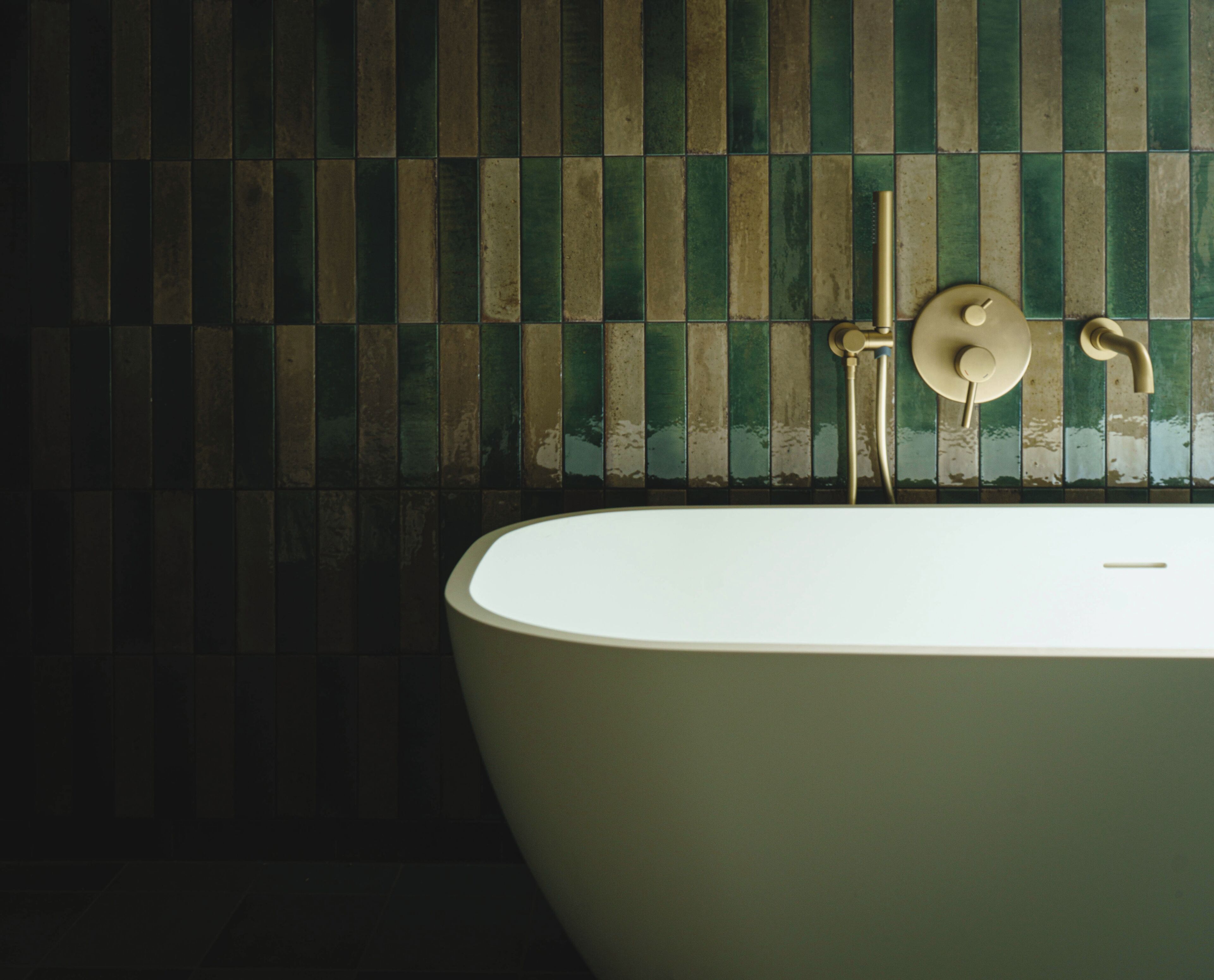 Une baignoire dotée d’un robinet et d’une pomme de douche dorés, placée devant un mur carrelé aux motifs verts et beiges.