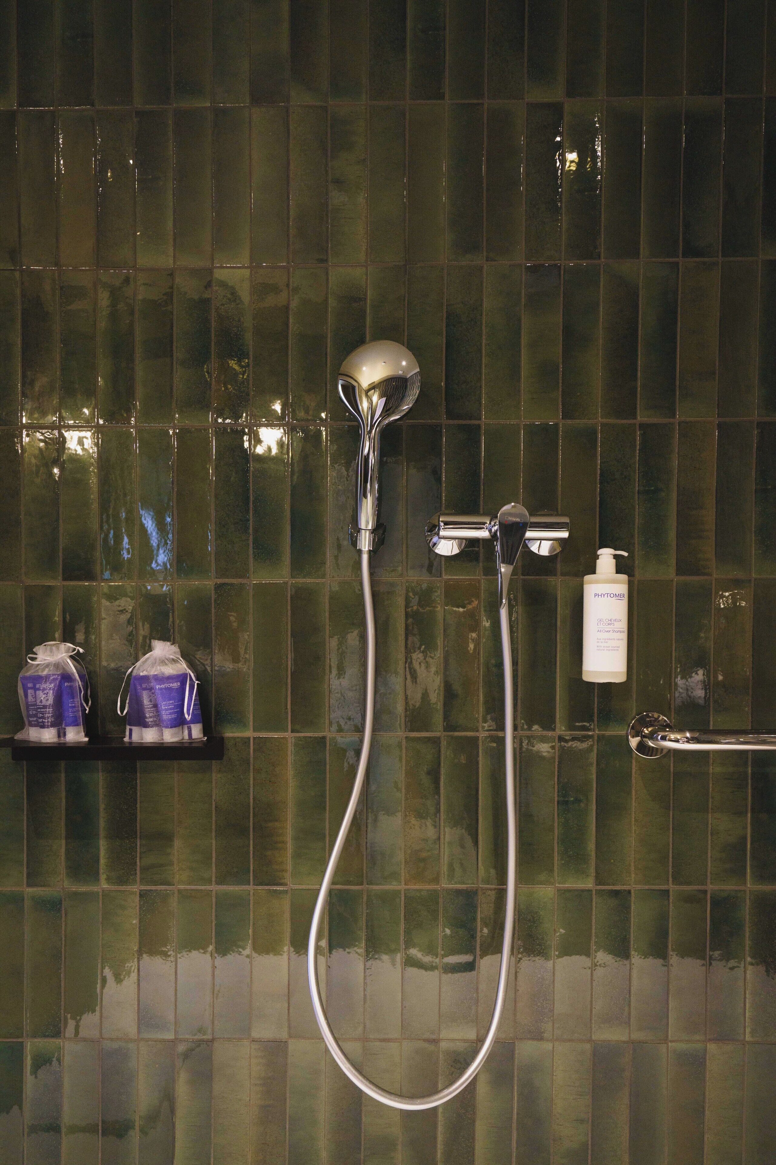 Ducha con cabezal de ducha manual y grifo empotrado en la pared, una botella de jabón Phytomenon y dos botellas de champú sobre un estante.