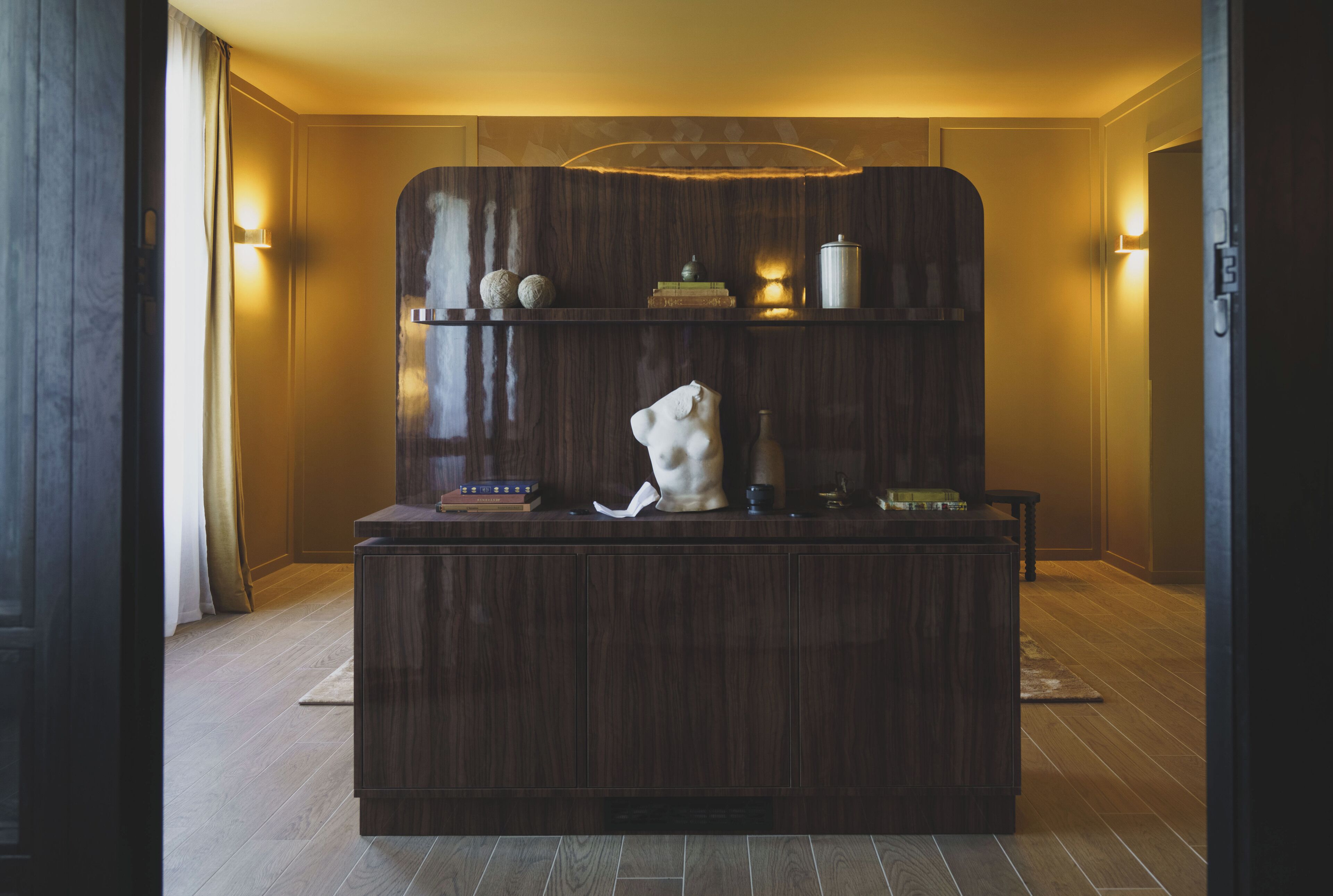 A reception desk with a wooden finish, a white bust sculpture, and a shelf with books and decorative items.