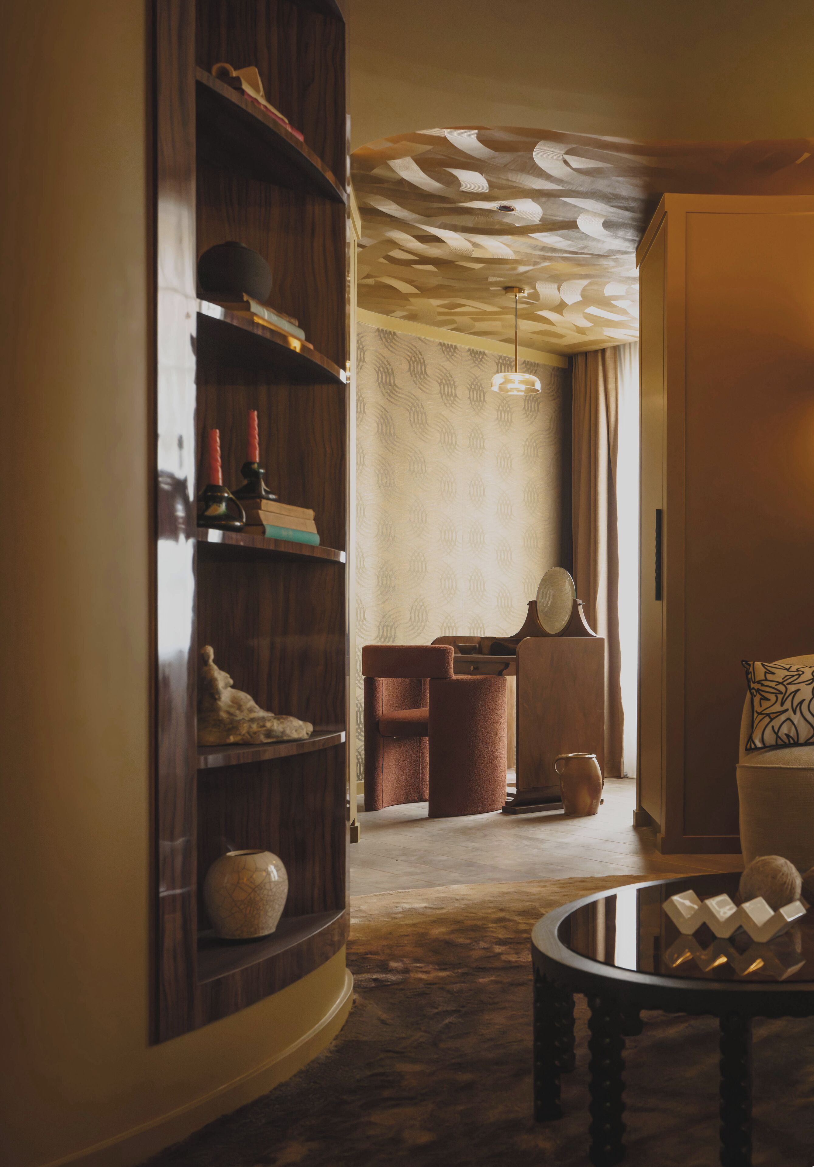 A room with a wooden bookshelf, a round table with decorative items, a brown armchair, and a patterned ceiling.