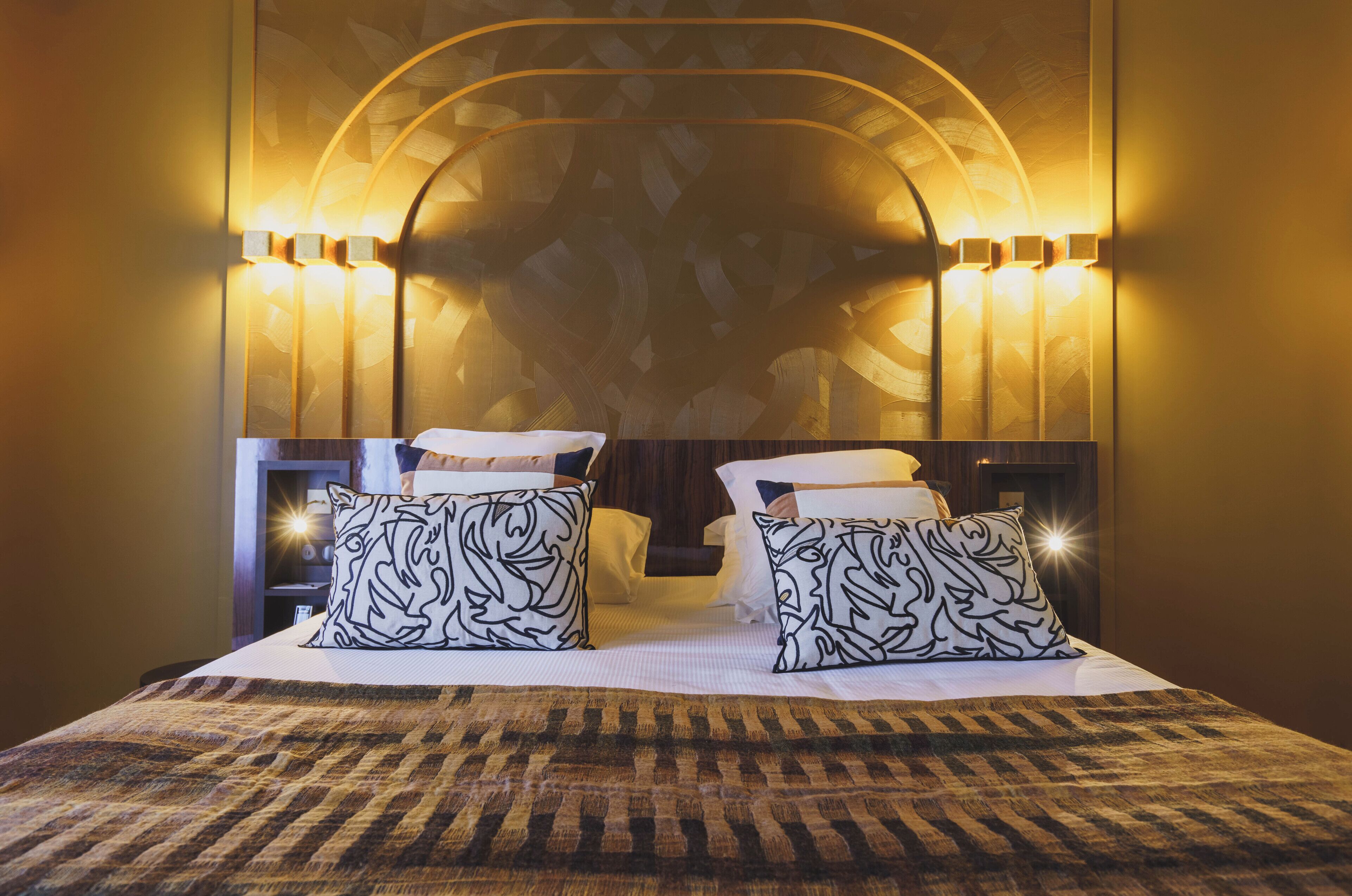 A hotel room with a large bed, two patterned pillows, a headboard with wall sconces, and a patterned rug.