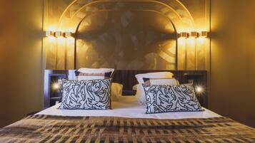 Habitación de hotel con una cama grande, dos almohadas estampadas, cabecera con apliques de pared y alfombra con diseño.