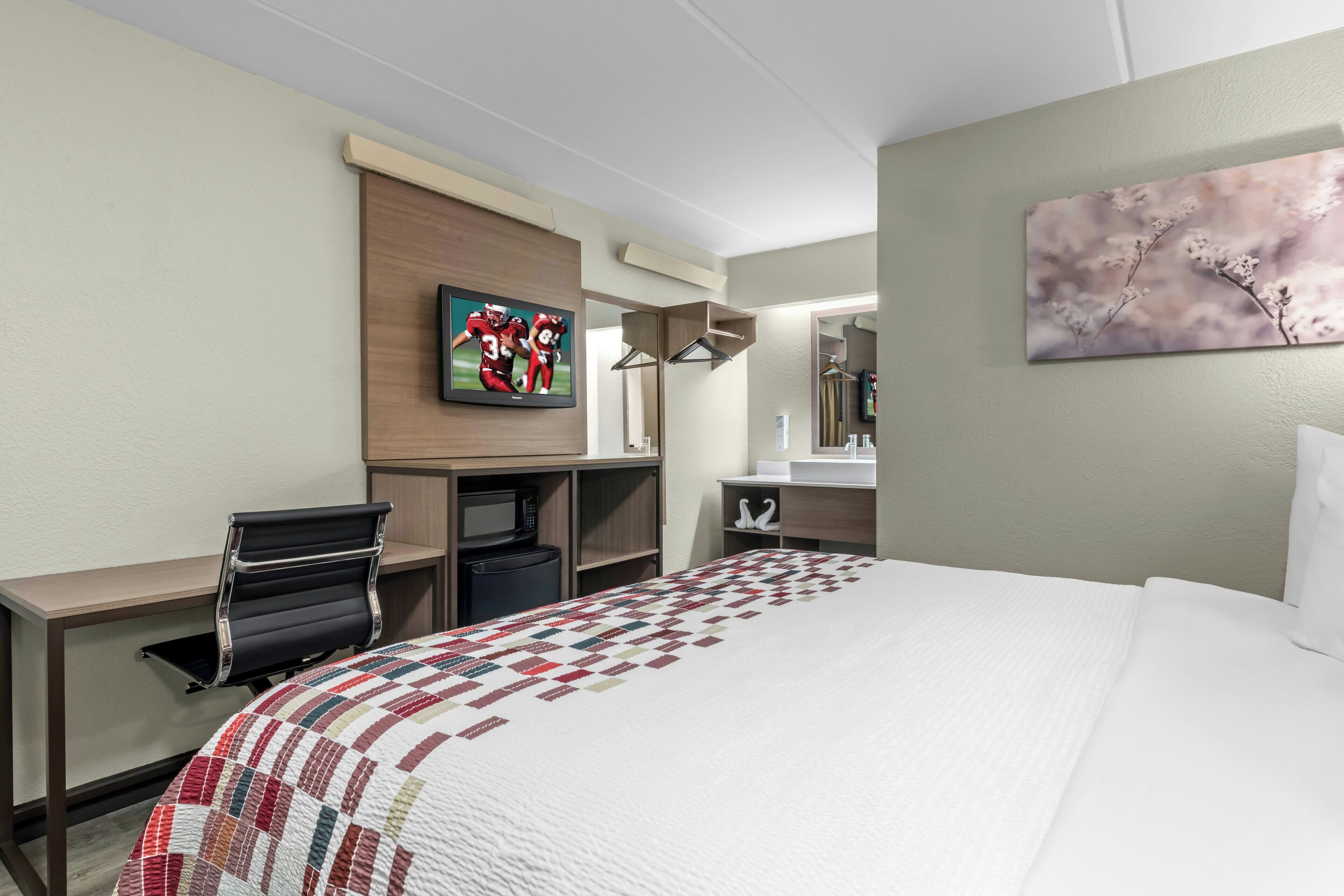 Habitación de hotel con cama, escritorio, televisión, microondas, refrigerador, espejo y un cuadro en la pared.