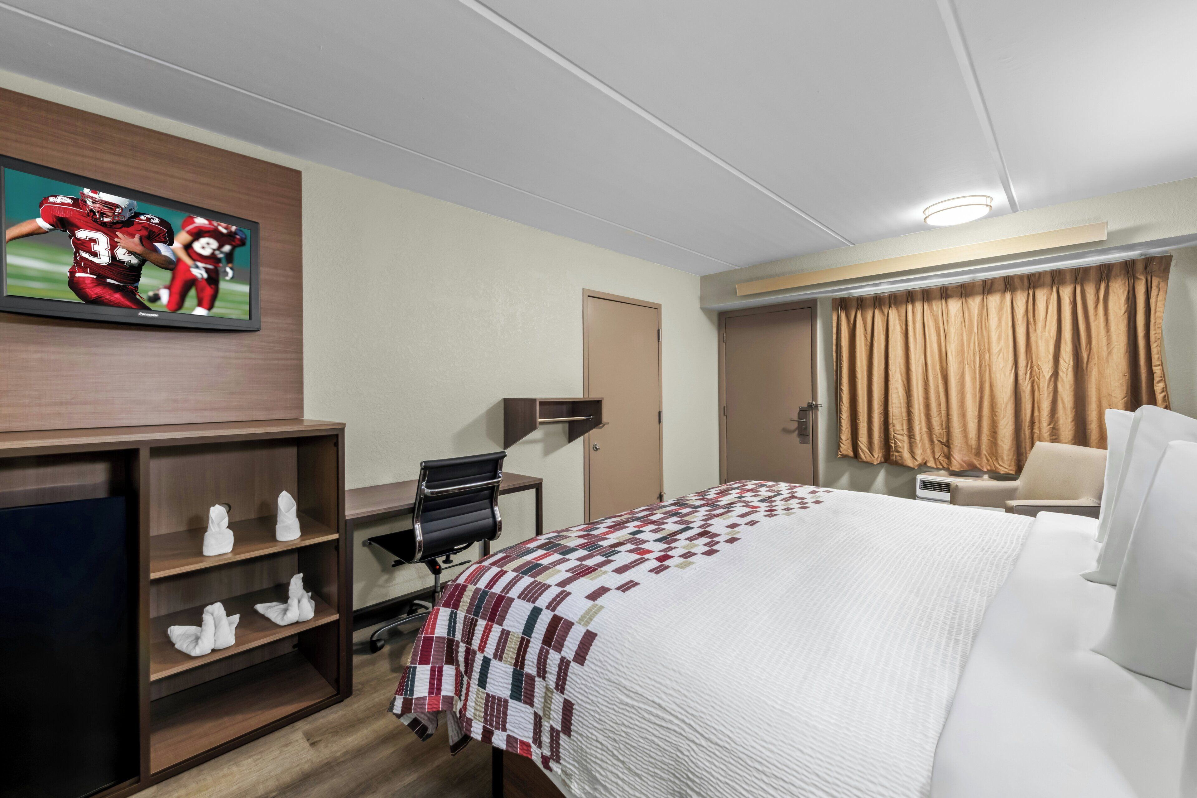 Hotellihuone, jossa on sänky, työpöytä, tuoli, televisio ja ikkuna, jossa on verhot.