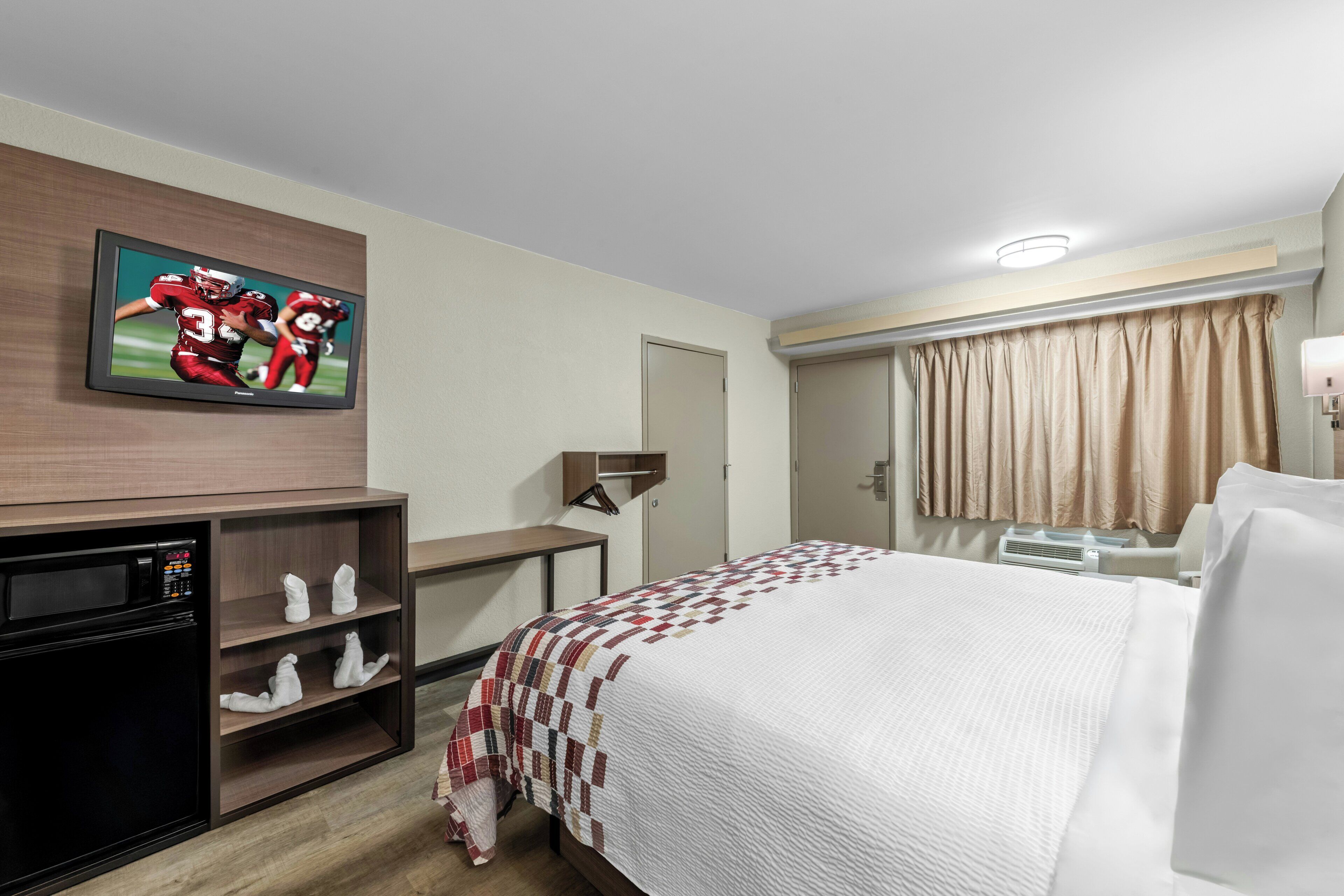 A hotel room with a bed, a TV mounted on the wall, a desk, a microwave, and a wardrobe.