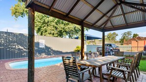 Um pátio externo com piscina, mesas e cadeiras de madeira, e uma área de estar coberta.