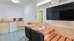 Cozinha compacta com micro-ondas, chaleira e pequena geladeira. Área de refeições com mesa posta para dois, incluindo pratos, copos e talheres. Uma TV de tela plana está fixada na parede acima da mesa.