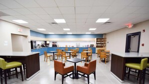 A spacious cafeteria with multiple tables and chairs, a counter with a coffee machine, and a blue and white color scheme.