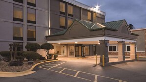 A hotel entrance with a covered walkway, a parking area, and a landscaped garden with shrubs and a small tree.