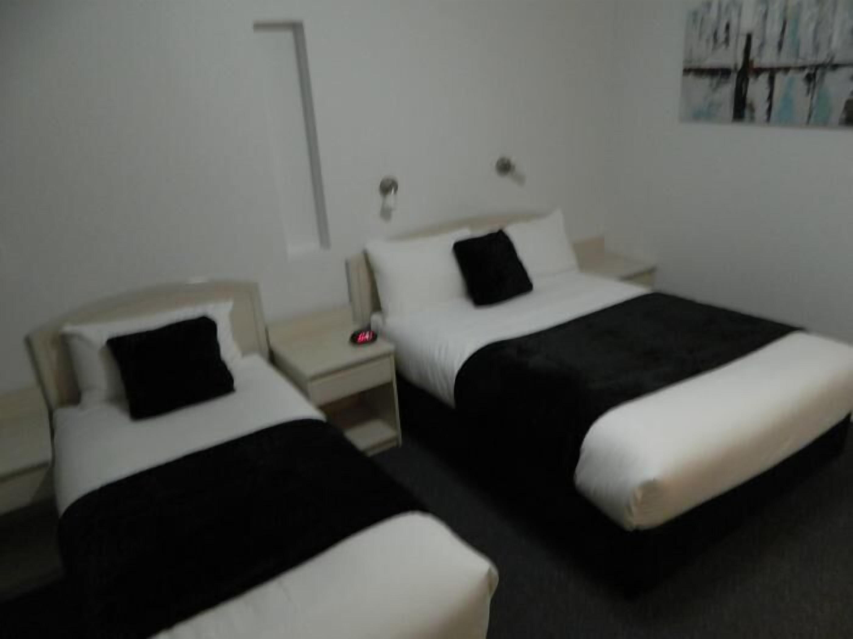 Habitación de hotel con dos camas, cada una con almohada negra y ropa de cama blanca. Entre las camas, una mesita de noche con un despertador rojo.
