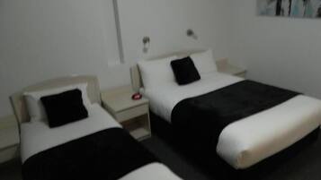 Habitación de hotel con dos camas, cada una con almohada negra y ropa de cama blanca. Entre las camas, una mesita de noche con un despertador rojo.