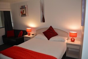 A hotel room with a bed, two nightstands, a red lamp, a red pillow, a black chair, and a wall art piece.