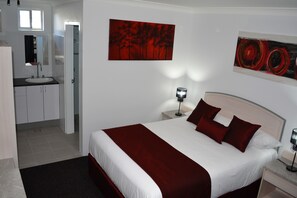 Una habitación de hotel con una cama grande, dos mesitas de noche, un tocador, una ventana, un baño con lavabo y espejo, y dos cuadros en la pared.