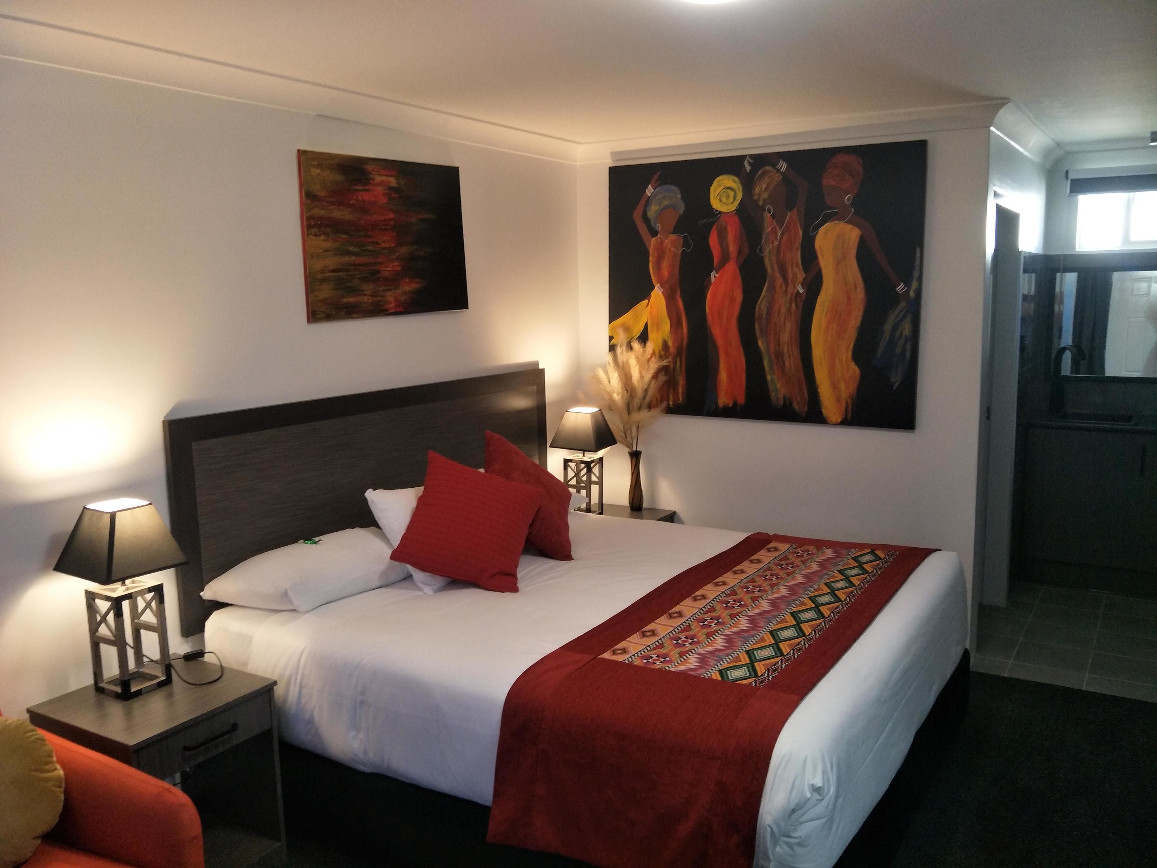 Una habitación de hotel con una cama grande, dos mesitas de noche con lámparas, una manta roja y dos cuadros en la pared.