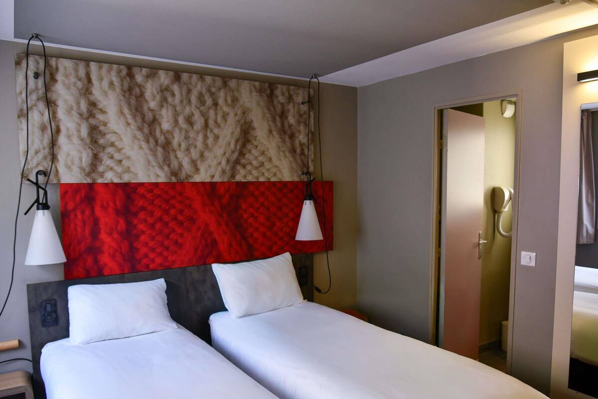 A hotel room with two single beds, a red and beige patterned headboard, a bedside table with a lamp, and a door leading to a bathroom.