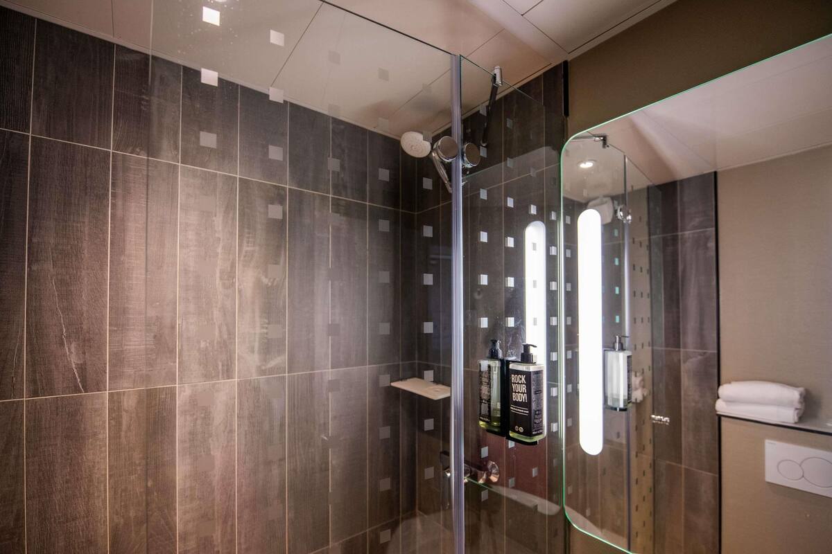 Un baño moderno con cabina de ducha de vidrio, paredes con azulejos oscuros y un espejo que refleja la habitación.