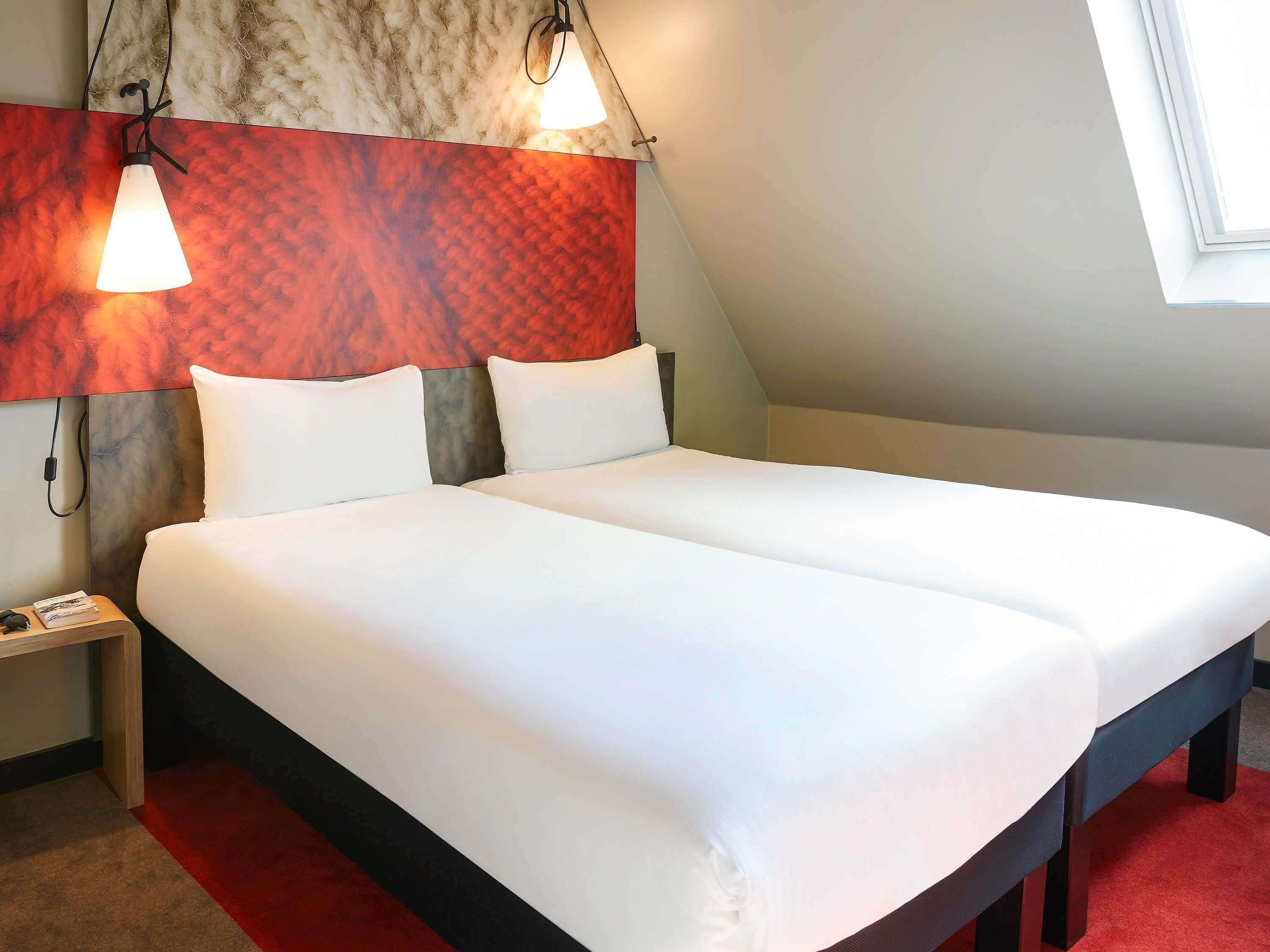 A hotel room with a large bed, a nightstand, a wall-mounted lamp, and a window.