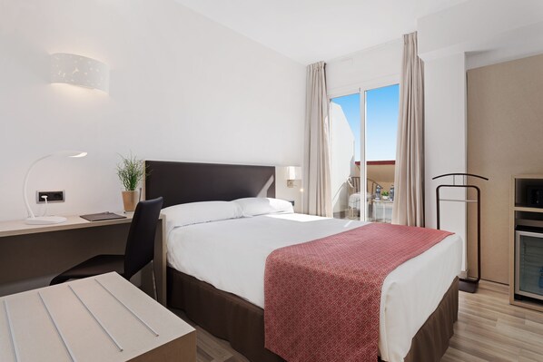 Une chambre d’hôtel moderne avec un grand lit, un bureau équipé d’un ordinateur, une chaise, une petite plante et une vue sur l’extérieur.
