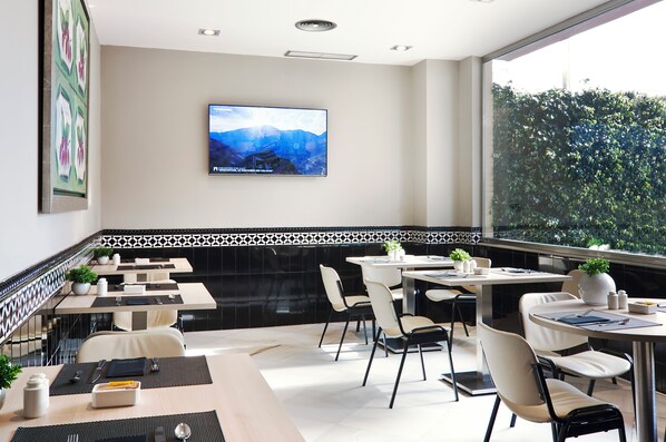 A modern restaurant with a large TV screen displaying a scenic view, black and white tiled backsplash, and a view of greenery through the window. - Catalonia Híspalis (Seville)