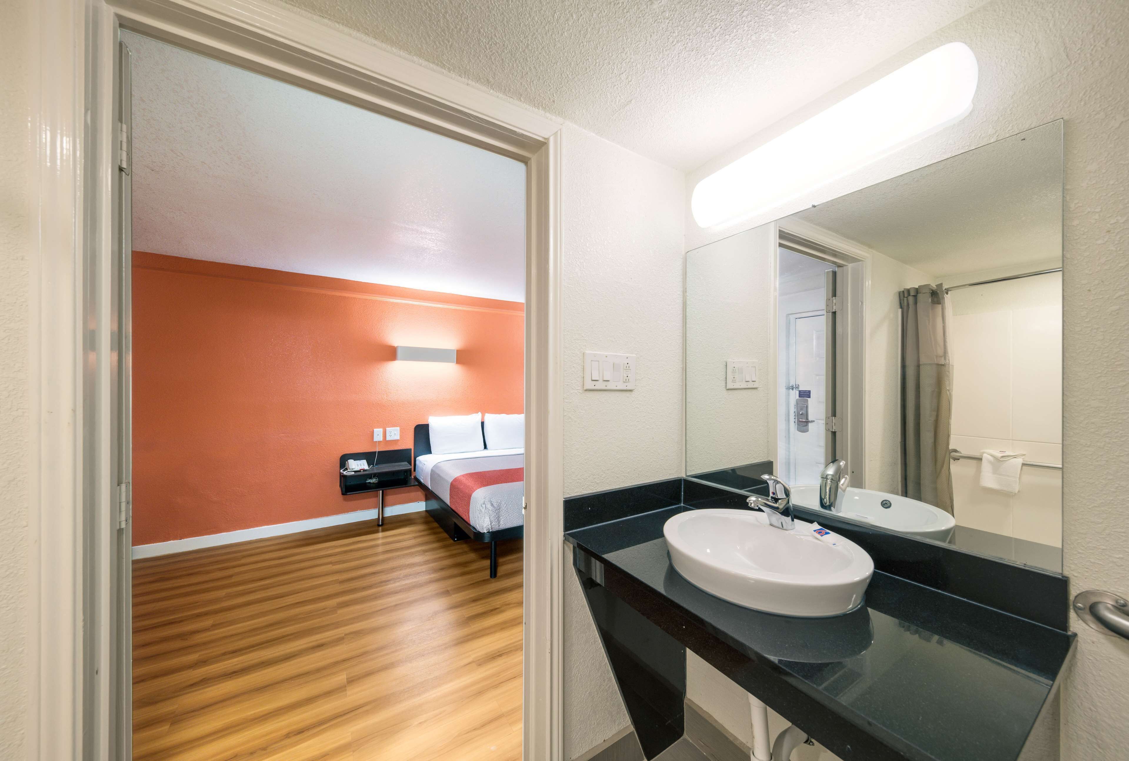 A hotel room with a wooden floor, a bed with white bedding, a black nightstand, a white sink, and a large mirror.
