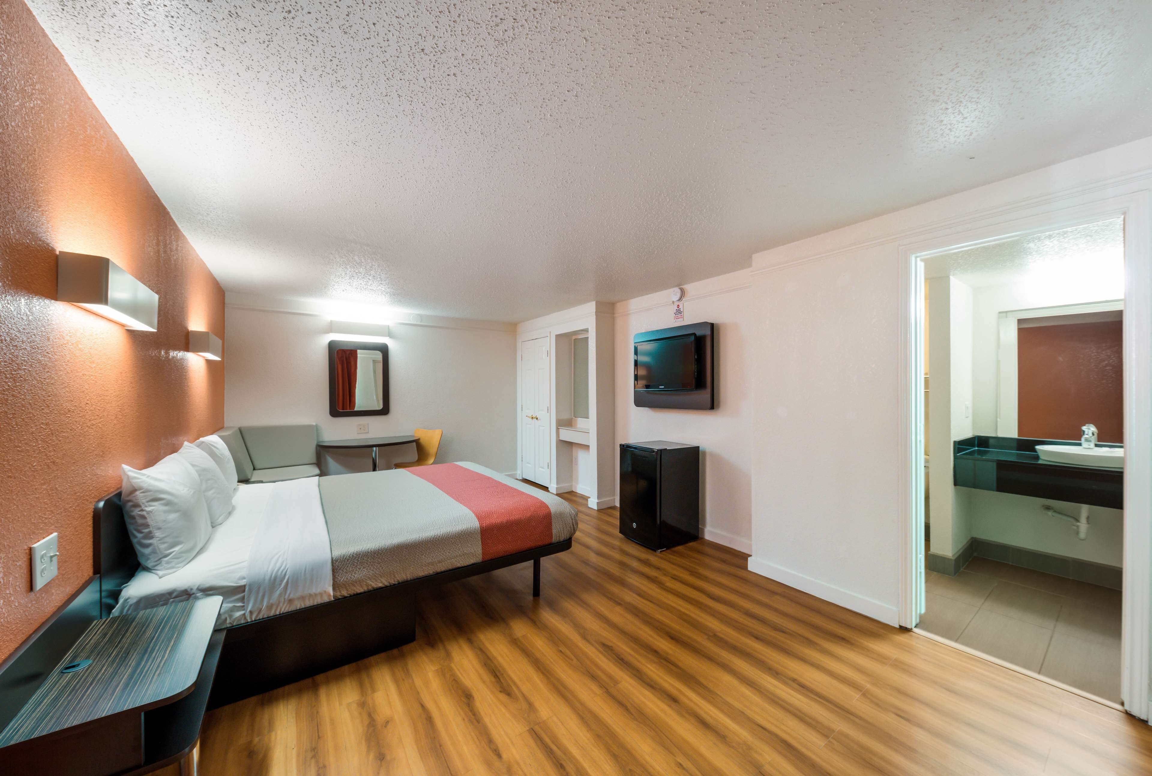 A modern hotel room with a wooden floor, a bed with a red and grey blanket, a desk with a lamp, a TV mounted on the wall, and a bathroom with a sink and mirror.