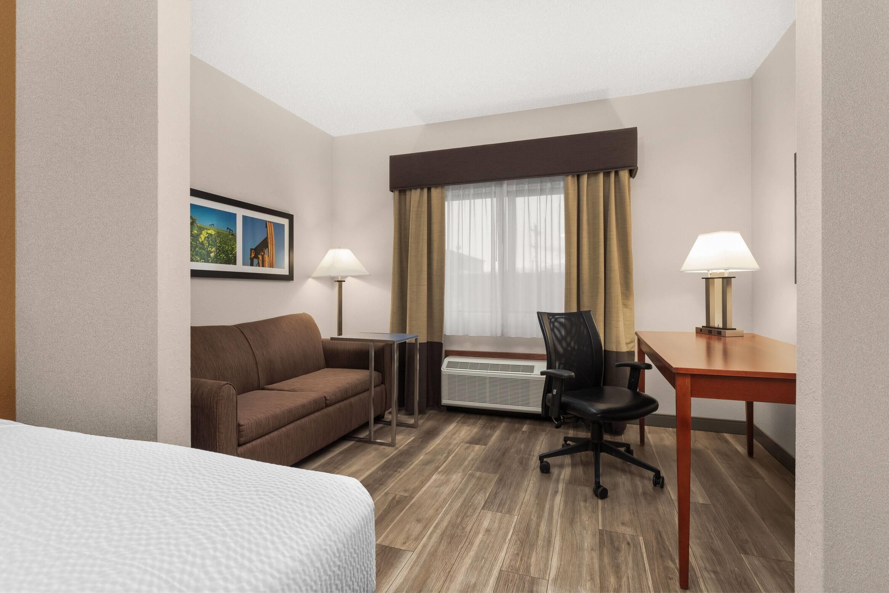 Habitación de hotel con cama, sofá, escritorio, silla, radiador y ventana con cortinas.