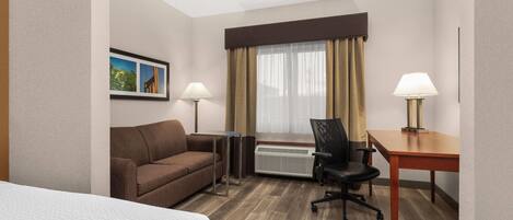 Habitación de hotel con cama, sofá, escritorio, silla, radiador y ventana con cortinas.