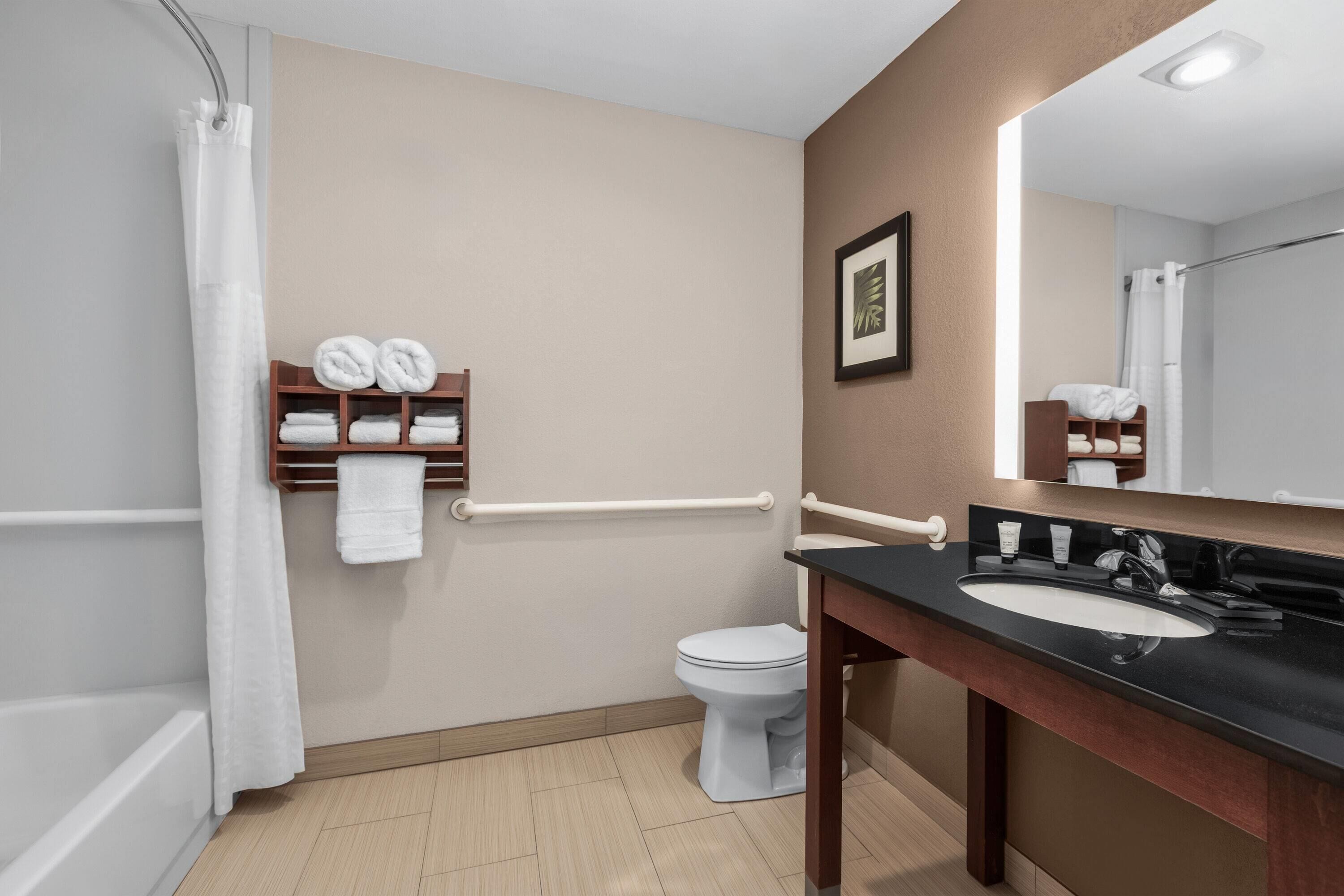 A bathroom with a shower, toilet, and sink. There are towels on a shelf and a framed picture on the wall.
