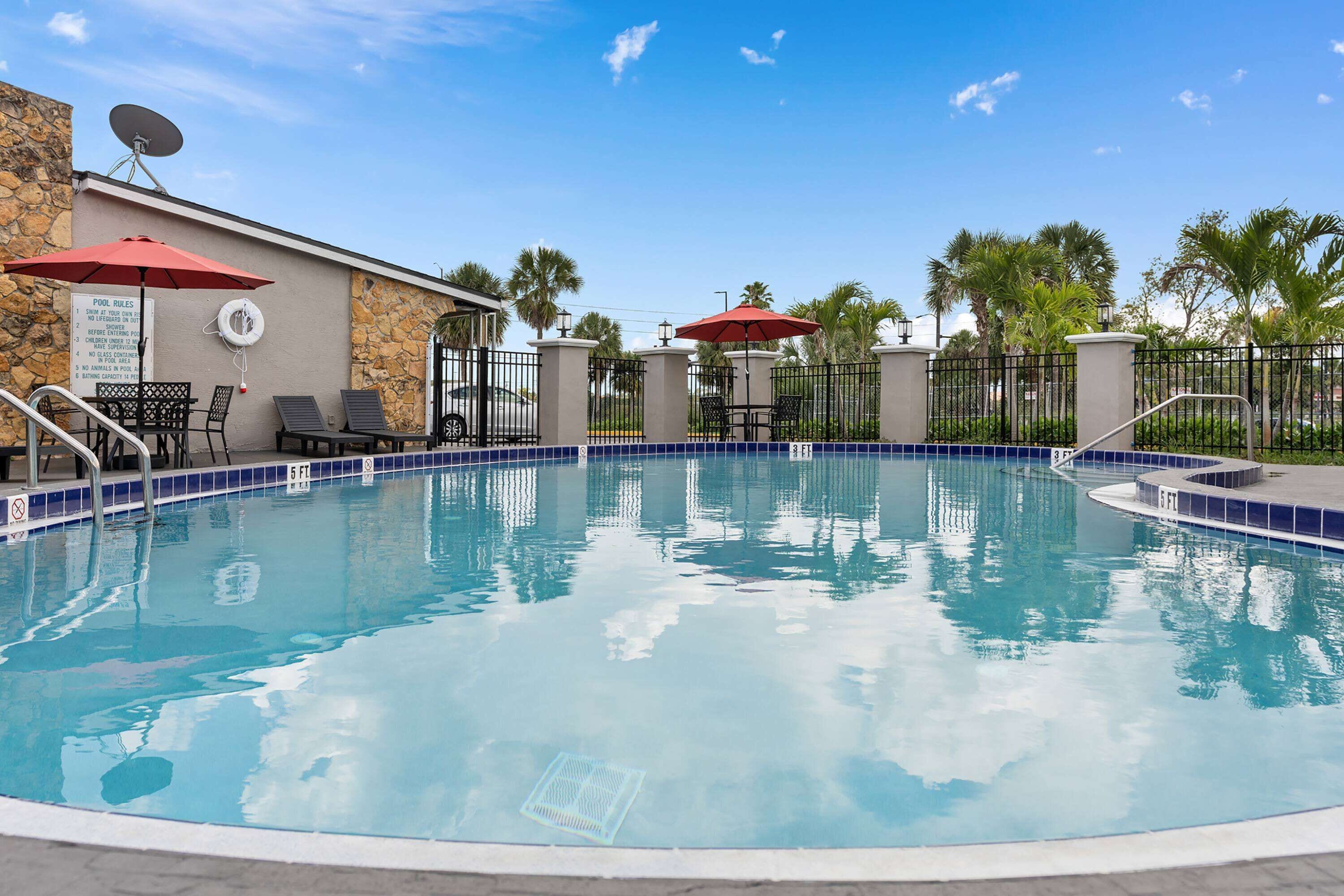 Una piscina con sillas de descanso y sombrillas, un cartel con las normas de la piscina y un muro de piedra.