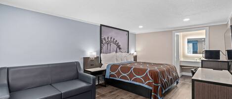 Una habitación de hotel con una cama, un sofá gris, un escritorio con una computadora y un cuadro en la pared.