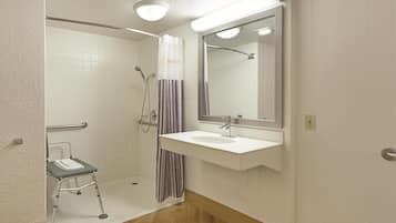 A bathroom with a shower, a sink, a mirror, and a wheelchair-accessible bench.