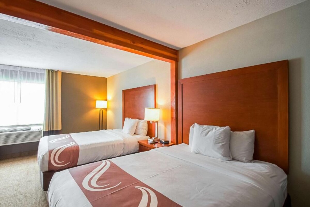 Quarto de hotel com duas camas, cabeceiras de madeira, mesa de cabeceira com abajur e uma janela com cortinas.