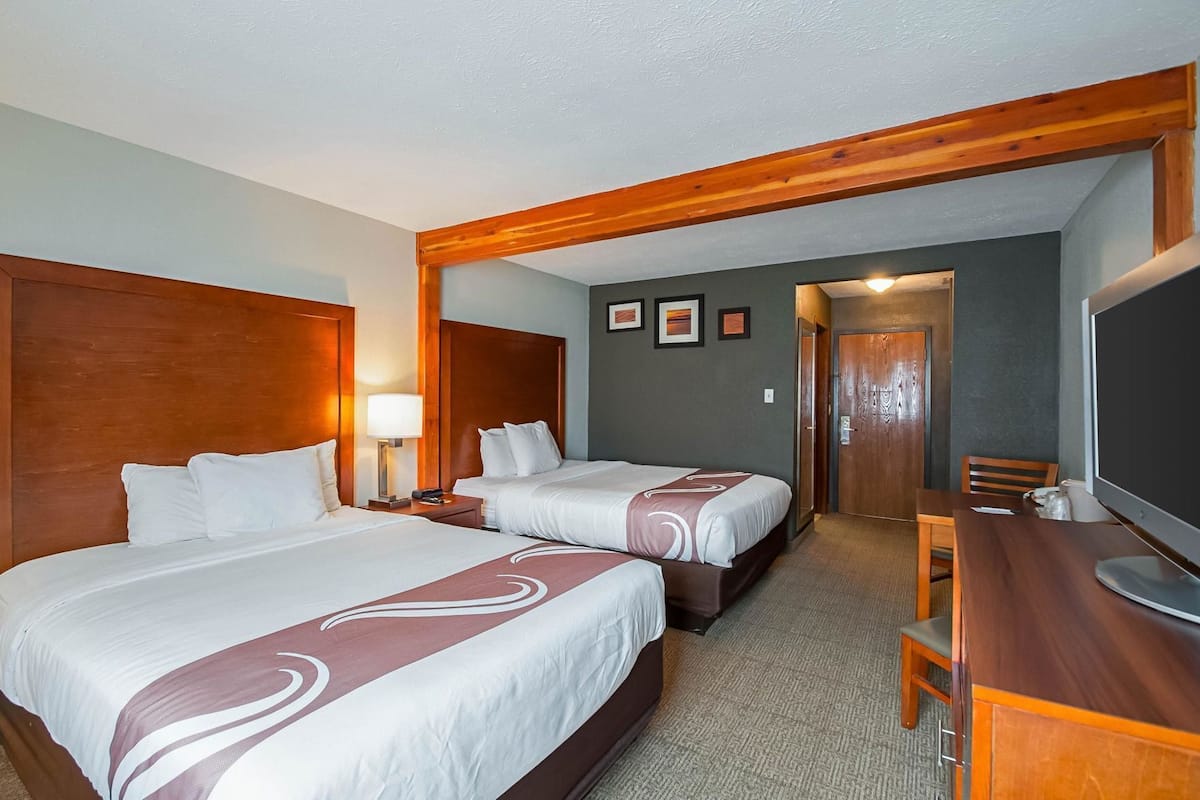Habitación de hotel con dos camas, un escritorio, una silla, un televisor y una puerta que da a un pasillo.