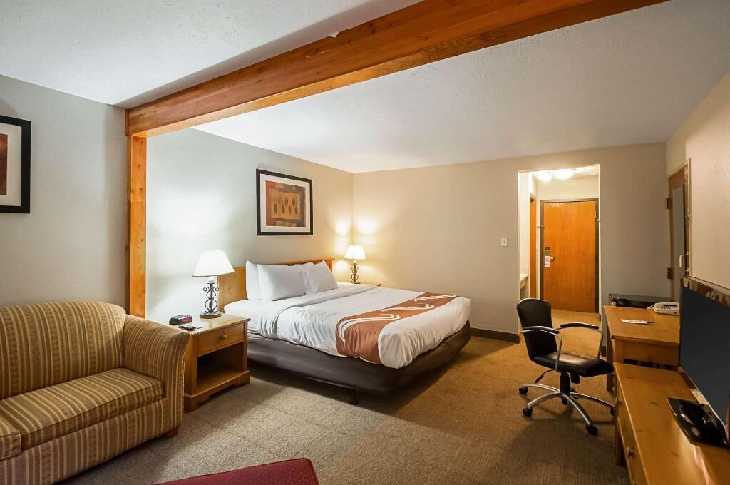A hotel room with a bed, a desk, a chair, a sofa, and a wooden ceiling beam.
