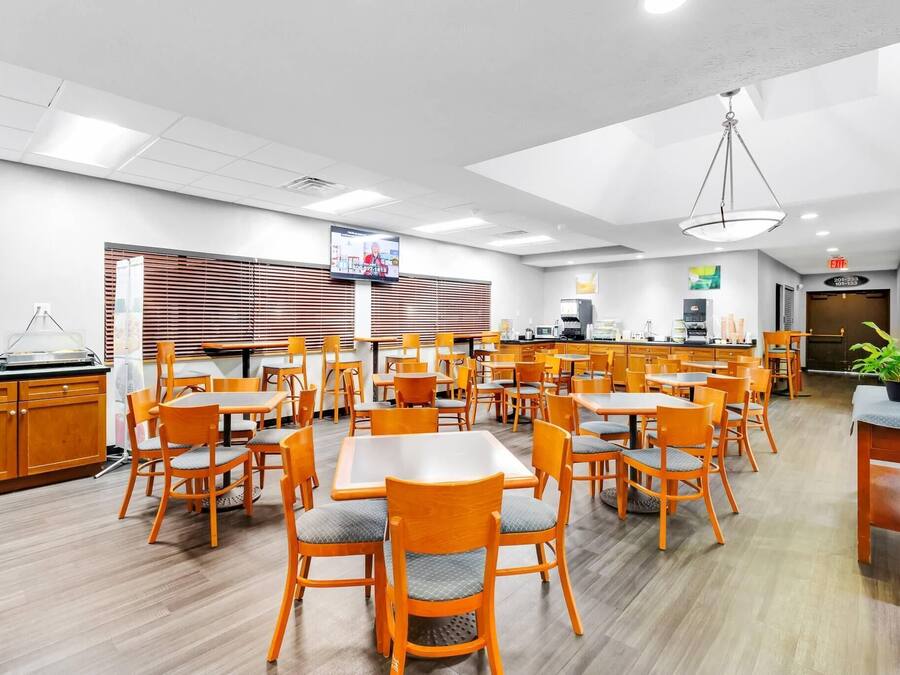 A spacious dining area with multiple tables and chairs, a kitchen in the background, and a television mounted on the wall.