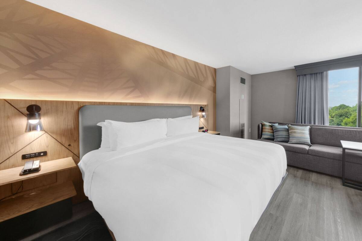 A modern hotel room with a large bed, a sofa, a nightstand, and a window with a view of greenery.