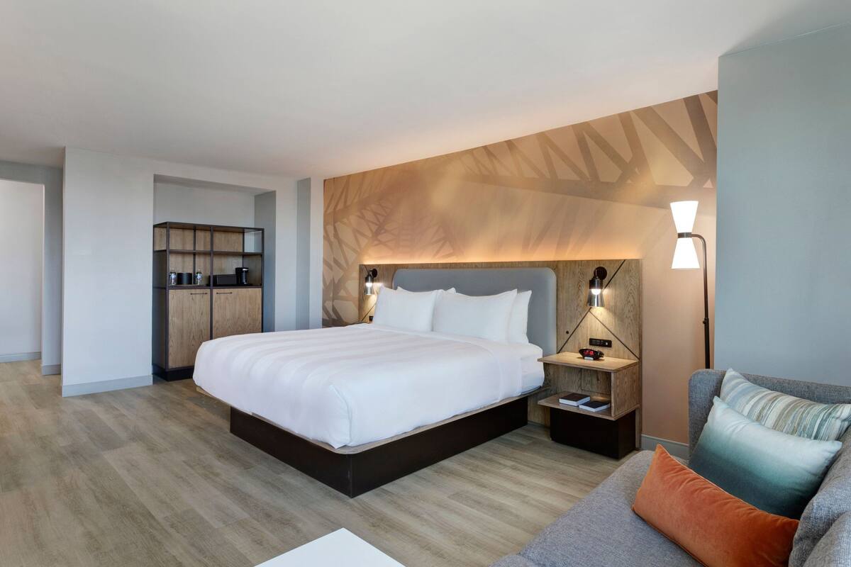 A modern hotel room with a large bed, a wooden headboard, a nightstand with a lamp, a sofa with cushions, and a built-in wardrobe.